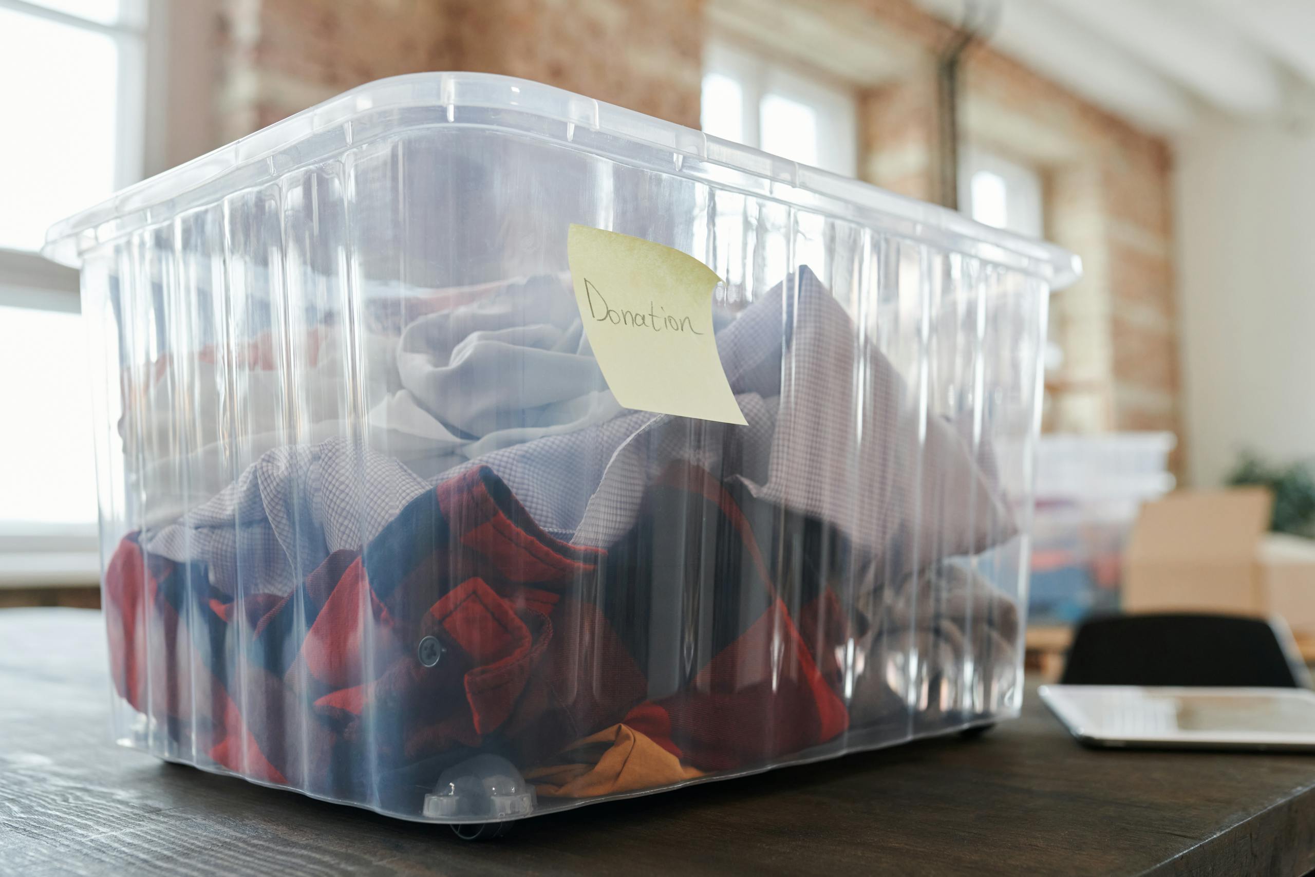 Clear plastic box filled with clothing for donation on a wooden table indoors, tagged donation.